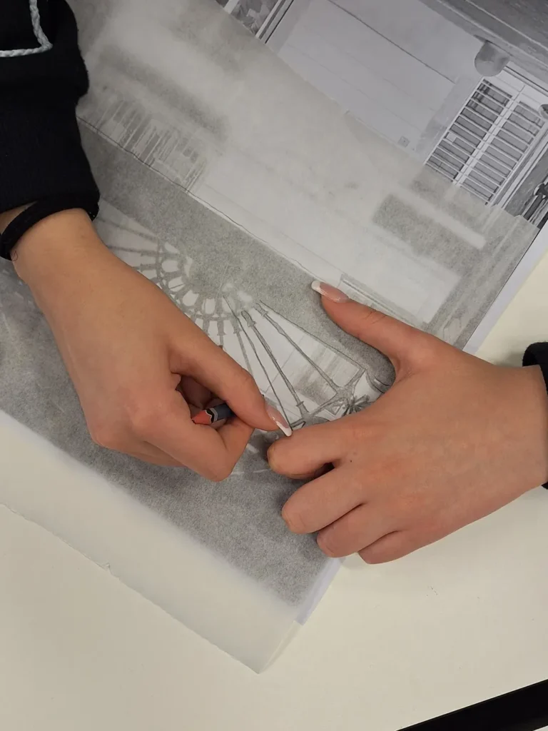 primo piano sulle mani di una studentessa che con l'uso di un foglio di carta trasparente ricalca da una foto della citta, delle forme utili al progetto